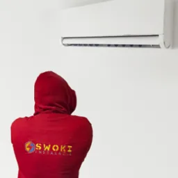 Mężczyzna w czerwonej bluzie z kapturem z logo firmy instalacyjnej stoi przed zamontowanym klimatyzatorem na białej ścianie.