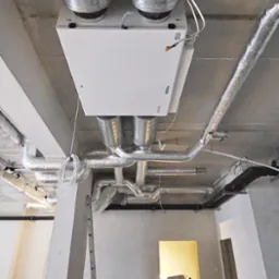 Centrala wentylacyjna z podłączonymi przewodami wentylacyjnymi, zamontowana pod betonowym stropem w surowym wnętrzu budynku w trakcie prac wykończeniowych.