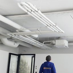 SWOKI - Białe jednostki klimatyzacyjne kasetonowe zamontowane na suficie biura, widoczne rury instalacyjne i pracownik w uniformie z logo firmy