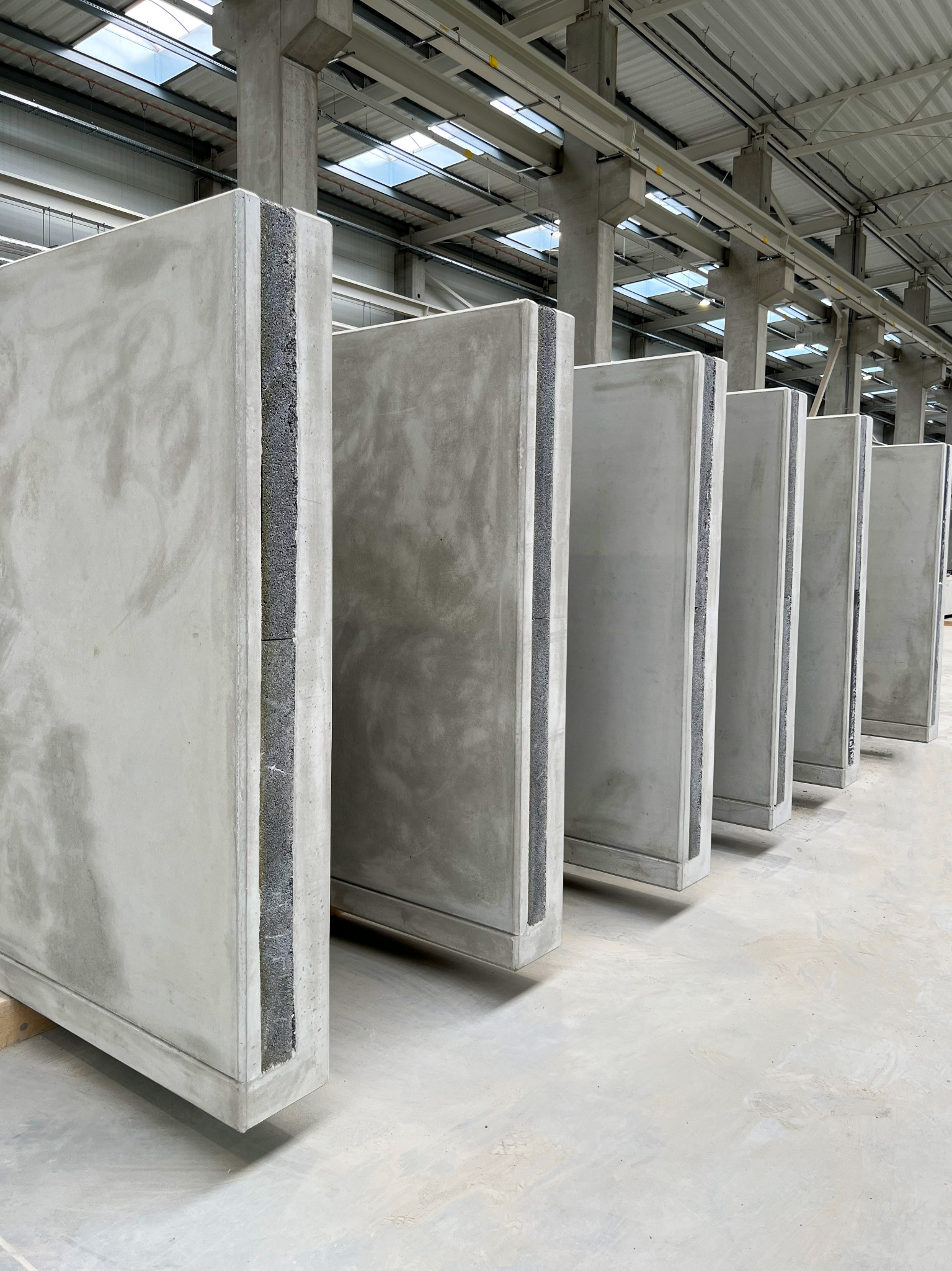 Rząd pionowo ustawionych, szarych paneli z izolacją w magazynie, widoczne słupy konstrukcyjne i świetliki dachowe.