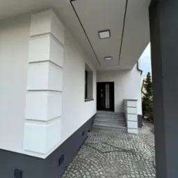 Nowoczesna elewacja domu z dekoracyjnymi elementami w postaci białych, kwadratowych bloków na rogu budynku, szara podmurówka i wejście wyłożone kostką brukową.