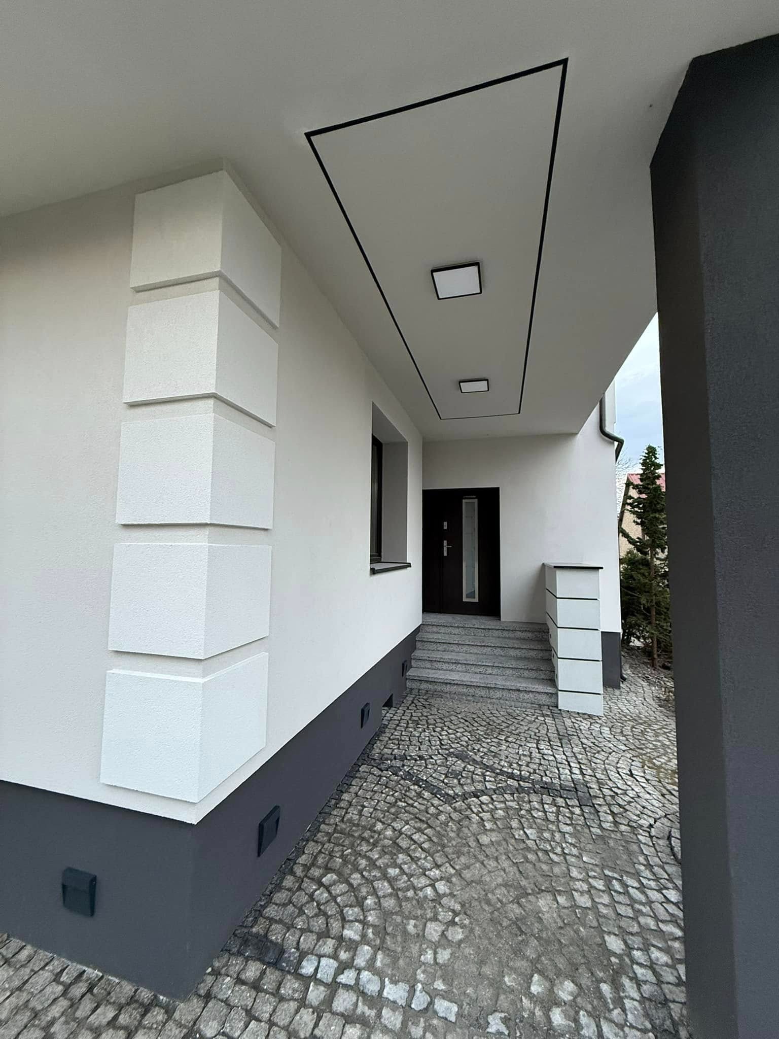 Nowoczesna elewacja domu z dekoracyjnymi elementami w postaci białych, kwadratowych bloków na rogu budynku, szara podmurówka i wejście wyłożone kostką brukową.