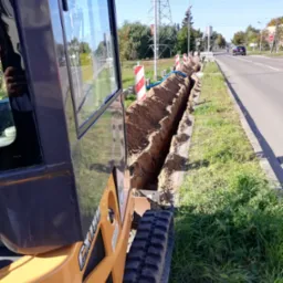 Mini koparka gąsienicowa obok wykopu wzdłuż drogi z widocznymi rurami instalacyjnymi i znakami ostrzegawczymi.