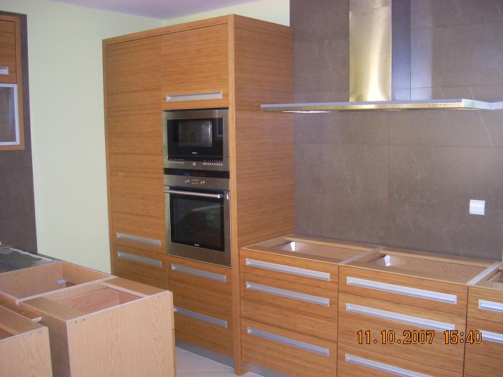 Montaż kuchni: szafki z drewnopodobną okleiną, zabudowa z piekarnikiem i mikrofalówką ze stali nierdzewnej, szare płytki na ścianie i okap, widoczne niedokończone szafki bez blatów.