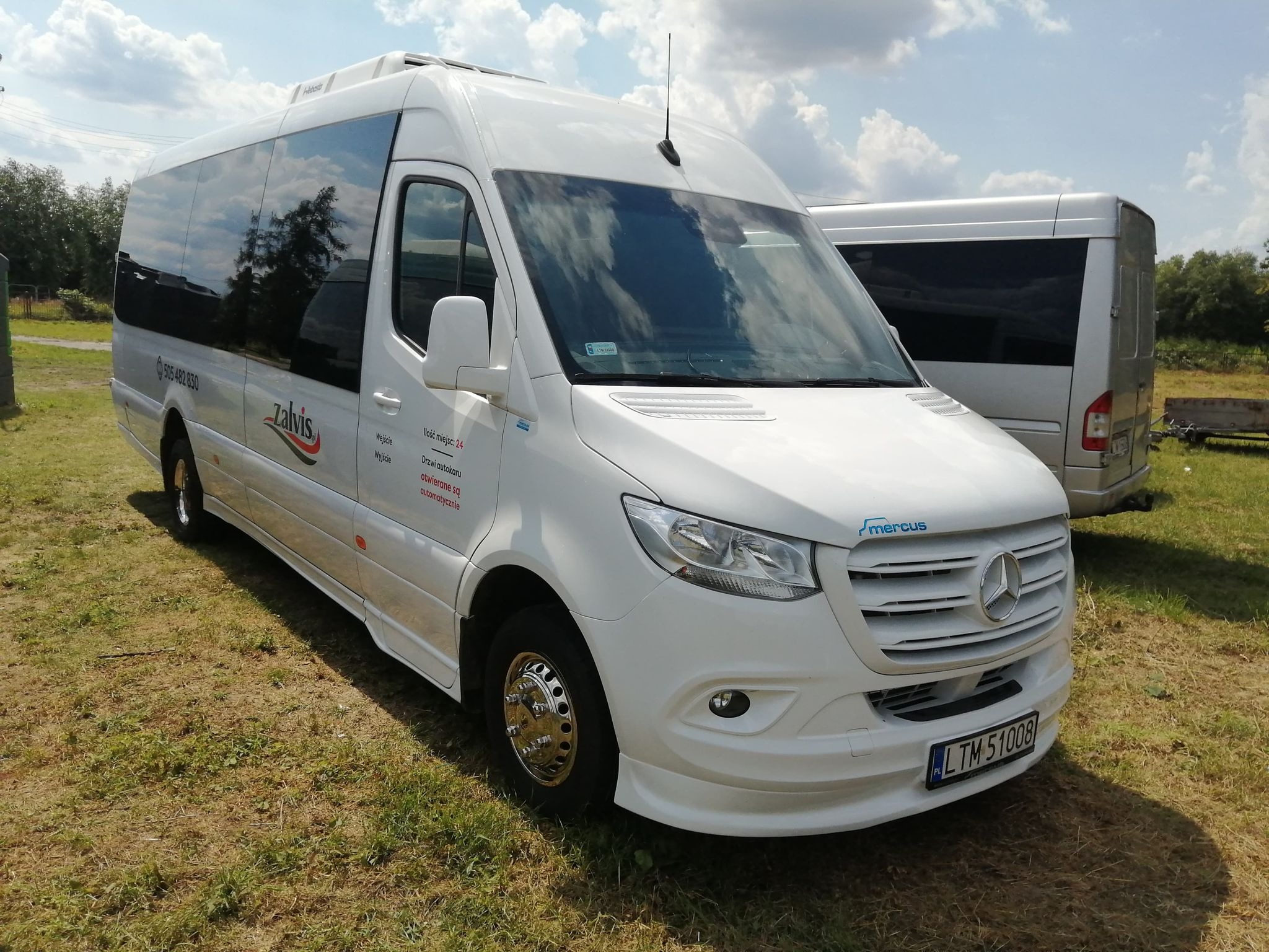 Biały bus Mercedes Sprinter z chromowanymi felgami na trawiastym terenie, widoczny numer rejestracyjny LTM 51008, logo 'zalvis' i 'mercus' oraz dane kontaktowe firmy.