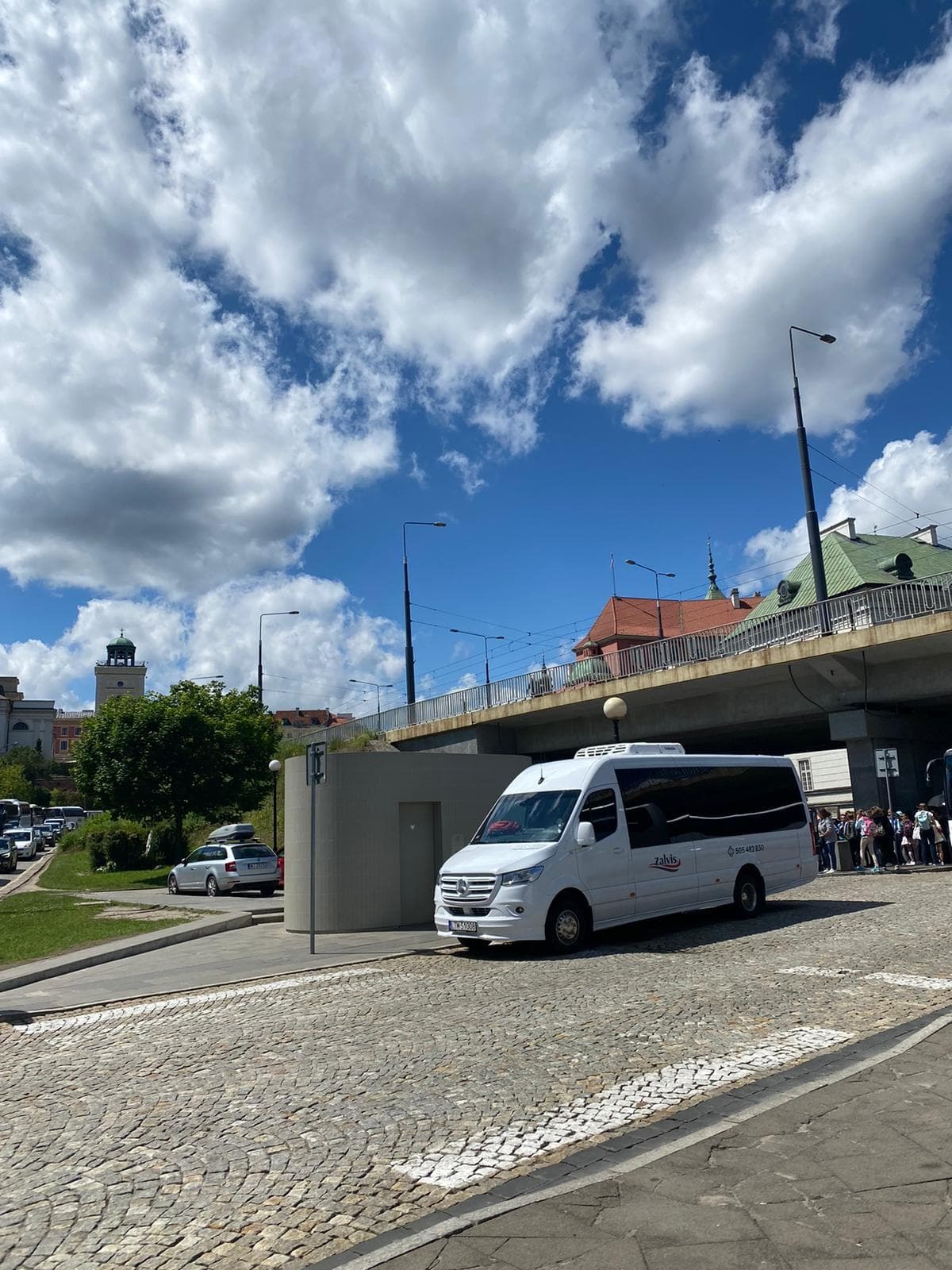 Biały bus marki Mercedes-Benz z czarnymi szybami, zaparkowany na brukowanej ulicy z widokiem na Stare Miasto w Warszawie, widoczny numer telefonu na karoserii i logo firmy przewozowej.