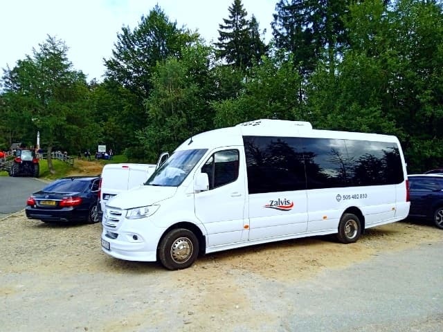 Biały bus marki Mercedes-Benz Sprinter z logo 'Zalvis' i numerem telefonu, zaparkowany na żwirowym parkingu z widokiem na drzewa i inne pojazdy w tle.