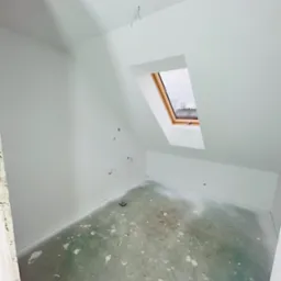 Poddasze z oknem dachowym w trakcie prac wykończeniowych; ściany pomalowane na biało, widoczne ślady farby na betonowej podłodze, puszki instalacyjne wystające ze ściany.