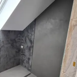 Wykończona łazienka na poddaszu ze ścianami pokrytymi szarą, dekoracyjną gładzią imitującą beton, widoczny fragment okna dachowego i odpływ liniowy w podłodze.