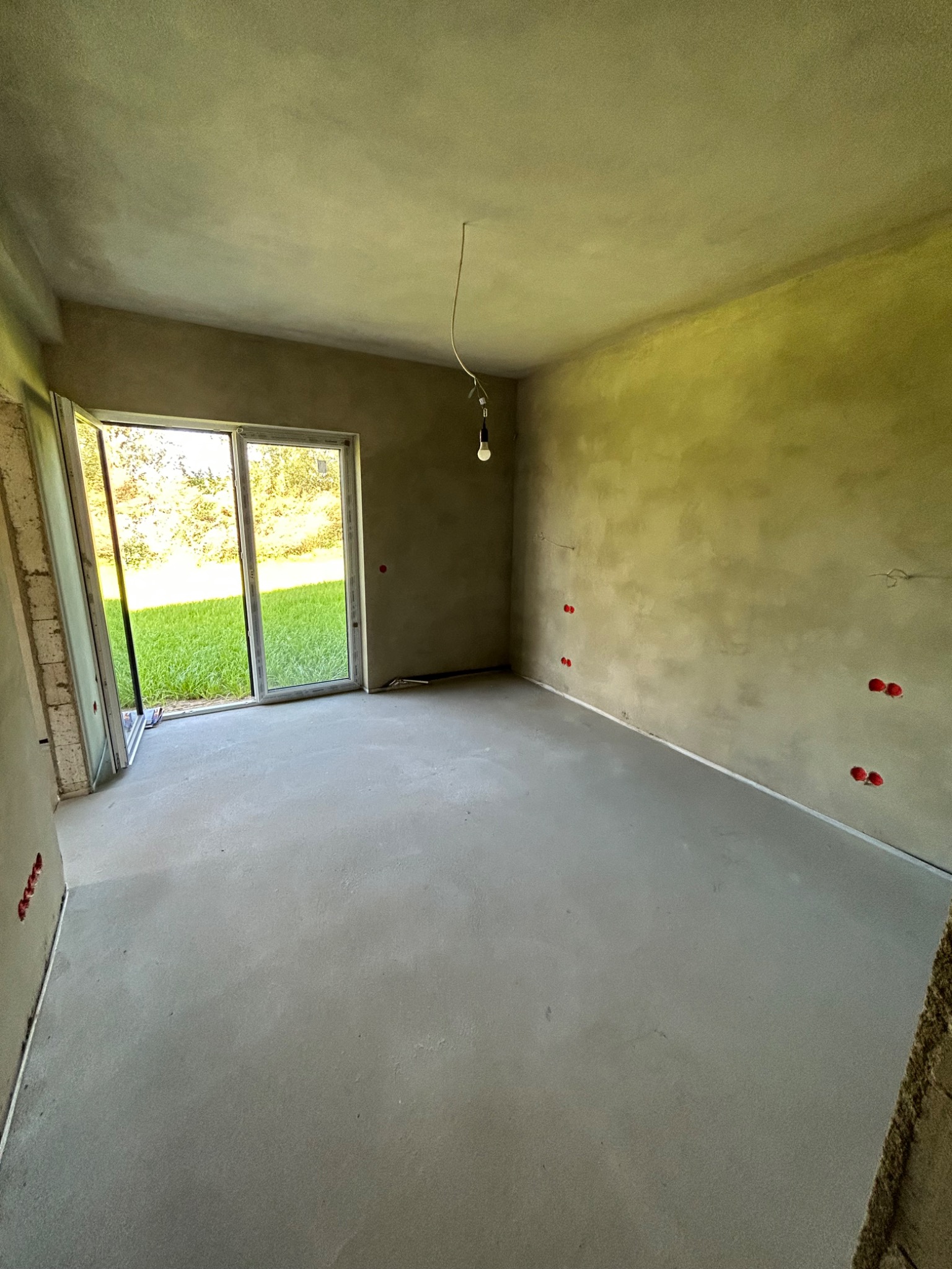 Pokój w trakcie prac wykończeniowych z nałożonymi tynkami, widoczne puszki elektryczne i surowa żarówka na kablu, z otwartym oknem balkonowym na zewnątrz z widokiem na trawnik.