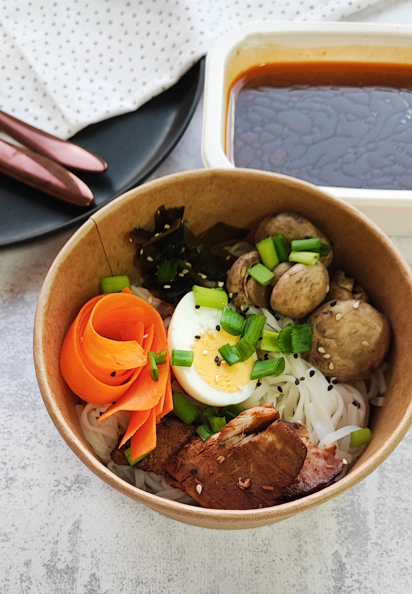 Porcja azjatyckiej zupy w papierowej misce: makaron ryżowy, jajko na twardo, pieczarki, wodorosty, marchewka w formie różyczki, mięso, posypane sezamem i zielonym szczypiorkiem. Obok sos w pojemniku.