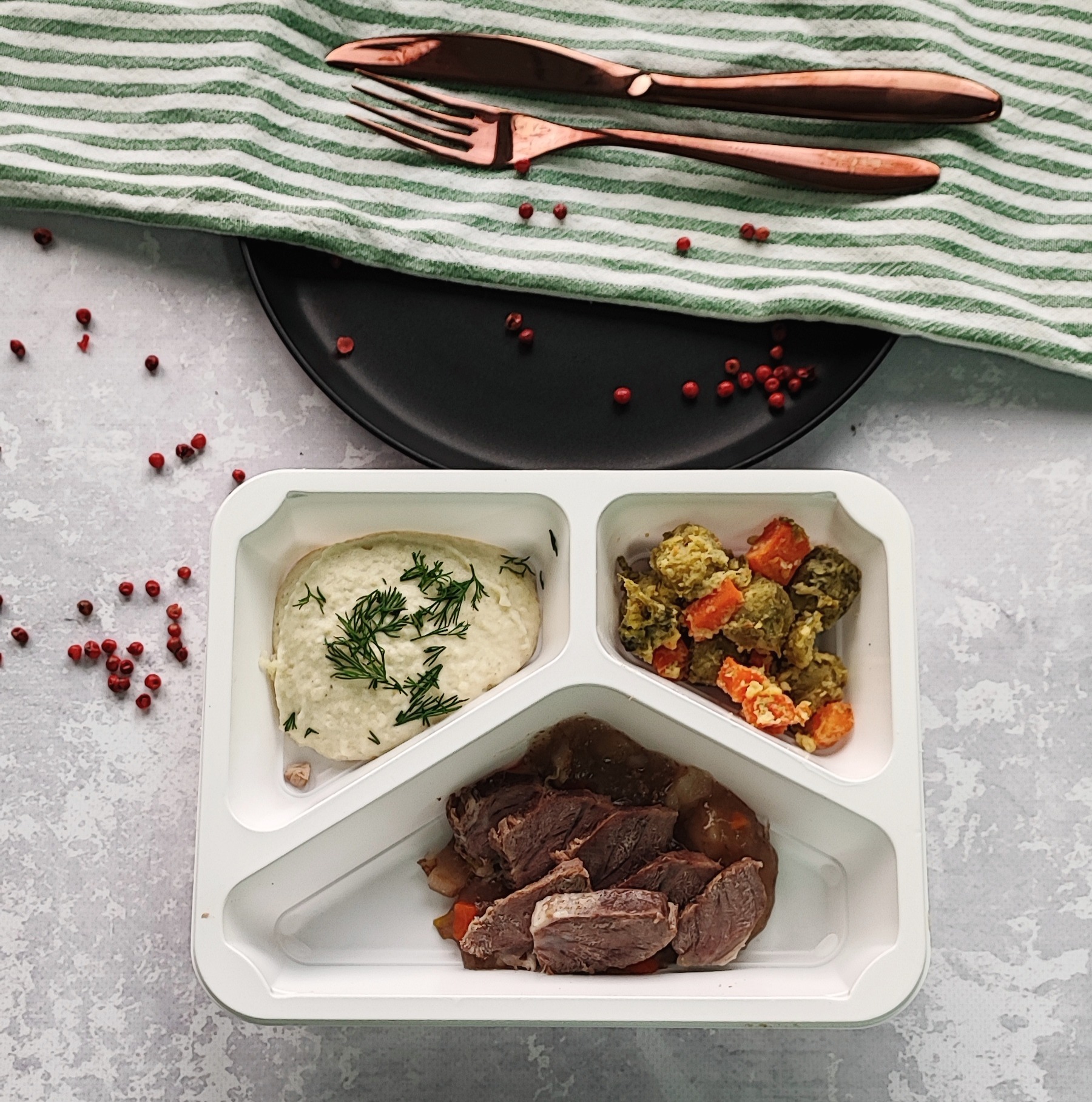 Gotowy zestaw posiłków w pudełku z trzema przegródkami, zawierający mięso, puree z koperkiem i warzywa, prezentowany na tle zielono-białej ściereczki, czarnego talerza i miedzianych sztućców,...