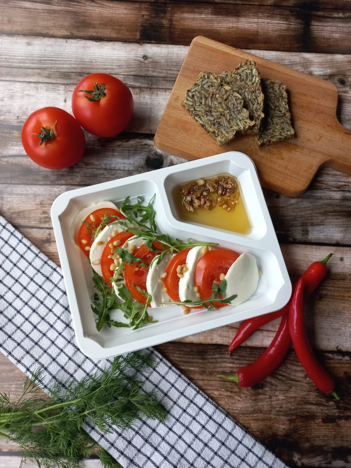 Dietetyczny posiłek w białym pudełku: Caprese z pomidorami, mozzarellą, rukolą i orzeszkami piniowymi, obok sos z orzechami, pieczywo z ziarnami na desce, pomidory, papryczki chili i koperek...