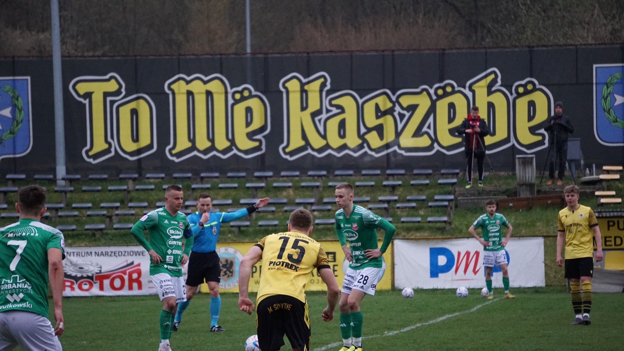 Mecz piłki nożnej: zawodnicy w zielonych i żółtych strojach na boisku, sędzia w niebieskim stroju, kibice na trybunach w tle, napis 'To Me Kaszebe' na czarnym banerze za trybunami.