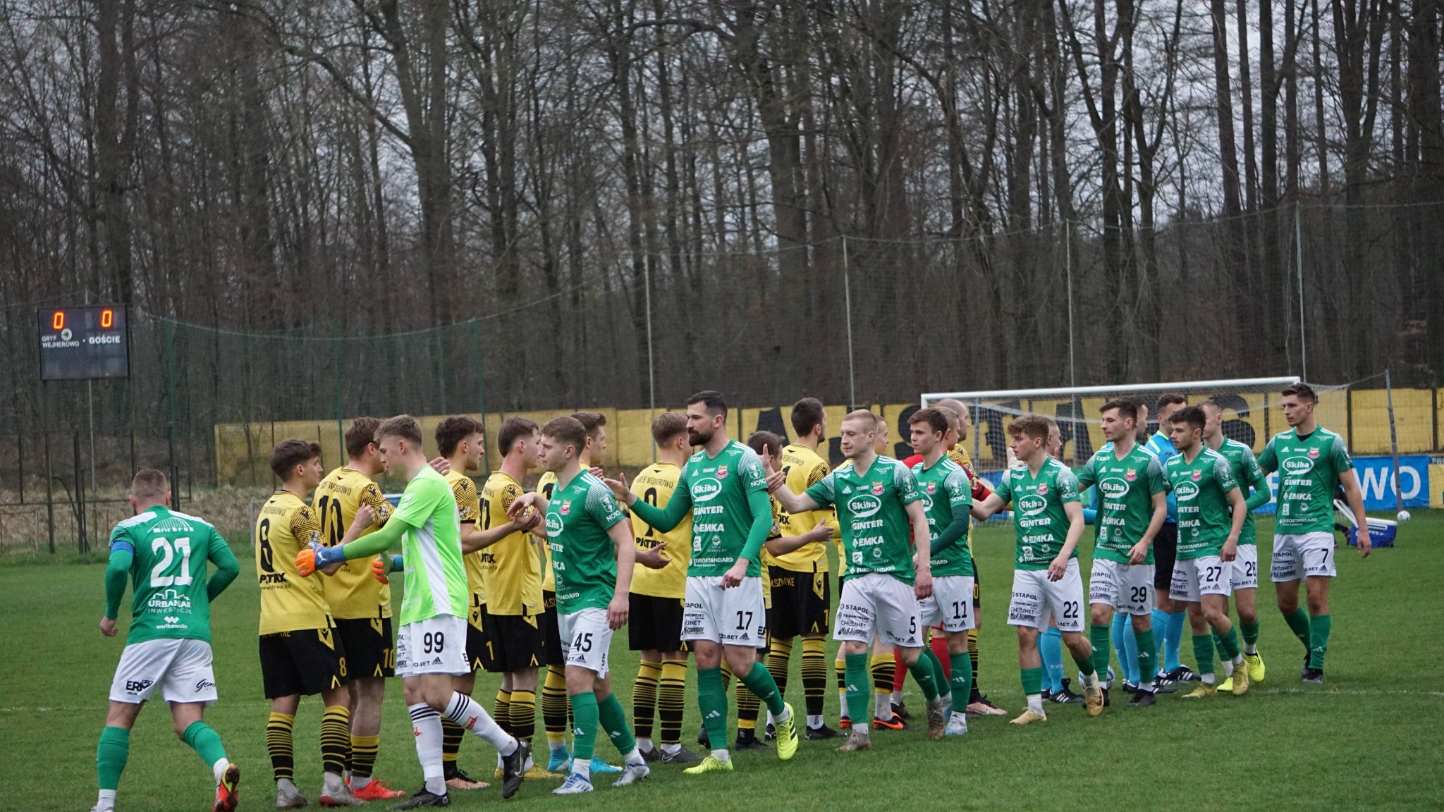 Piłkarze dwóch drużyn w strojach zielono-białych i żółto-czarnych witają się przed meczem na tle brązowych drzew i tablicy wyników.