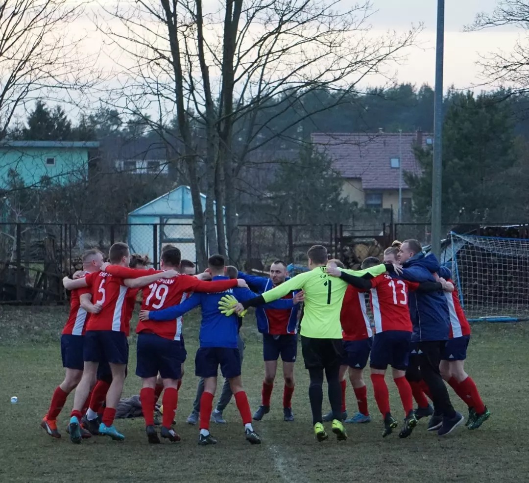 Drużyna piłkarska w czerwono-niebieskich strojach, obejmująca się w kręgu na boisku, pośród drzew i zabudowań w tle.