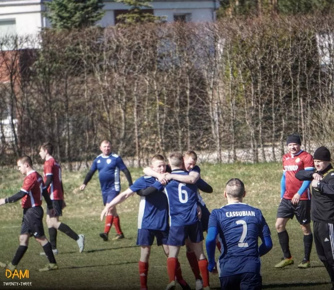 Grupa piłkarzy w niebieskich strojach świętuje zwycięstwo, obejmując się na boisku; w tle widoczni zawodnicy drużyny przeciwnej w czerwonych strojach i trener w czarnym dresie.