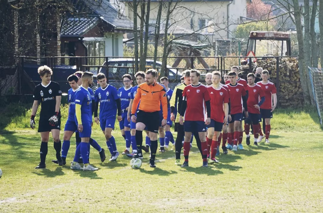 Dwie drużyny piłkarskie w strojach niebieskich i czerwonych, prowadzone przez sędziego w pomarańczowej koszulce, wchodzą na boisko z widocznym traktorem i stertą drewna w tle.