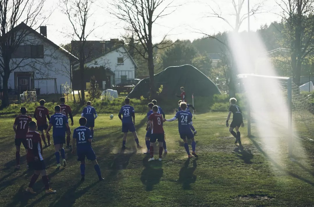 Mecz piłki nożnej amatorskiej na wiejskim boisku, zawodnicy w niebieskich i bordowych strojach w trakcie akcji, w tle domy i drzewa, silne promienie słoneczne.