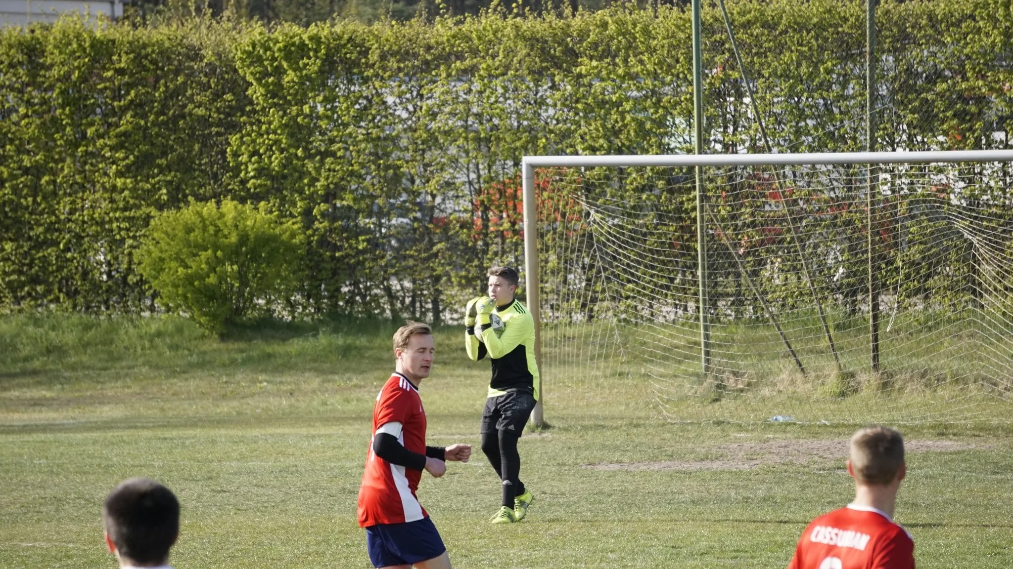 Bramkarz w jaskrawej koszulce ochrania bramkę podczas meczu piłki nożnej na trawiastym boisku, w tle żywopłot.