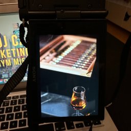 TLB ONLINE SPÓŁKA Z OGRANICZONĄ ODPOWIEDZIALNOŚCIĄ - Ekran aparatu Sony prezentuje zdjęcie cygar w pudełku i kieliszka z bursztynowym trunkiem, umieszczony na klawiaturze laptopa. Widoczny pasek aparatu i fragment ekranu laptopa.