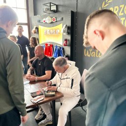 TLB ONLINE SPÓŁKA Z OGRANICZONĄ ODPOWIEDZIALNOŚCIĄ - Dwóch mężczyzn podpisuje zdjęcia przy drewnianym stole w pomieszczeniu z bokserskimi akcentami i widzami. Jeden z nich ma tatuaże na szyi i rękach.