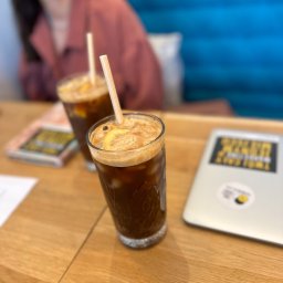 TLB ONLINE SPÓŁKA Z OGRANICZONĄ ODPOWIEDZIALNOŚCIĄ - Dwa drinki z lodem i cytryną na drewnianym stole obok laptopa z naklejką. W tle rozmyta postać kobiety i niebieska kanapa. Ujęcie z bliska, naturalne światło.