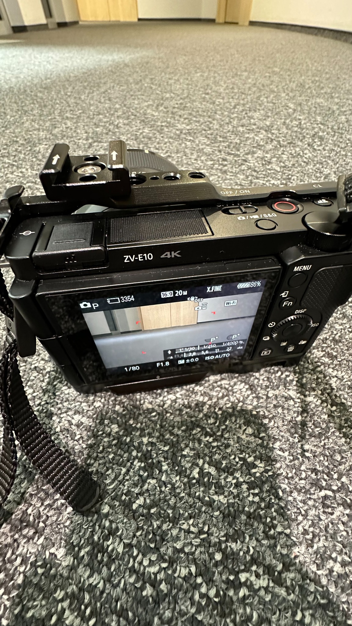 Aparat Sony ZV-E10 4K leżący na szarym dywanie. Widoczny ekran LCD z ustawieniami ekspozycji: 1/80s, f/1.8, ISO Auto. Ustawienia aparatu widoczne na ekranie.