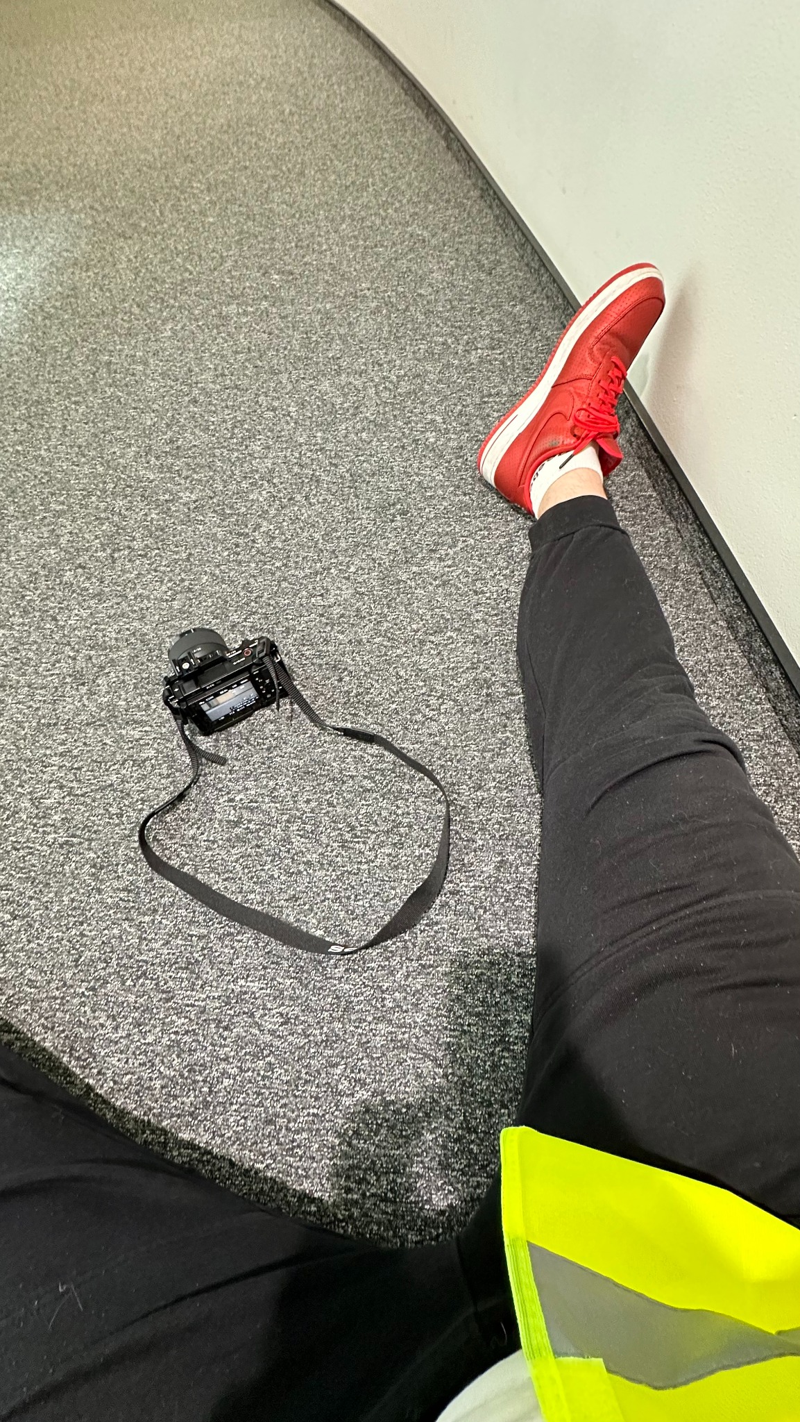 Aparat fotograficzny leżący na podłodze obok nogi w czerwonym sneakersie, widok z góry, w tle fragment ściany. Nietypowe ujęcie fotografa.
