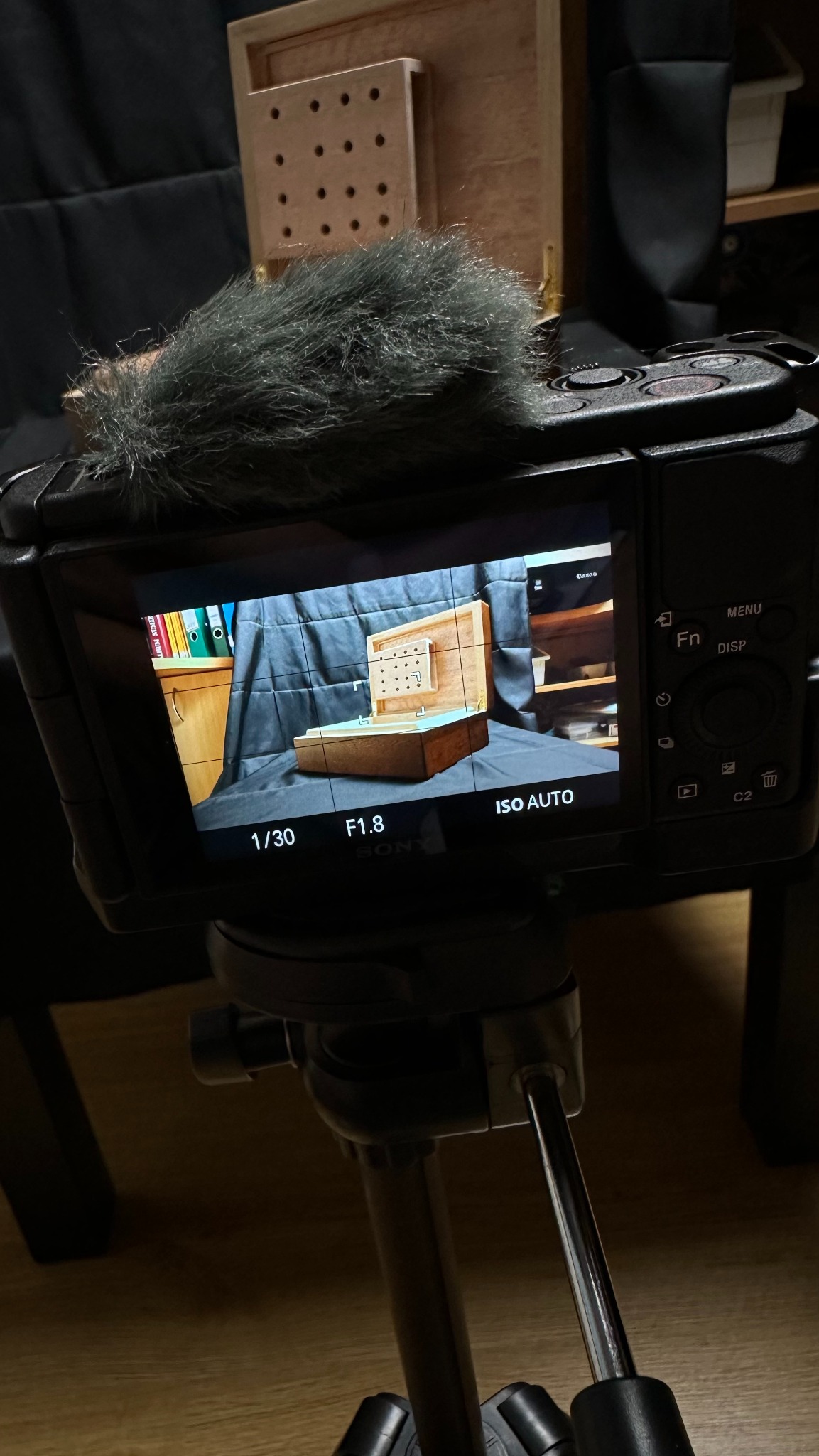 Ekran aparatu na statywie pokazuje drewniane pudełko z perforowaną płytką w środku, ustawione na czarnym tle. Widoczne ustawienia aparatu: 1/30, F1.8, ISO AUTO.