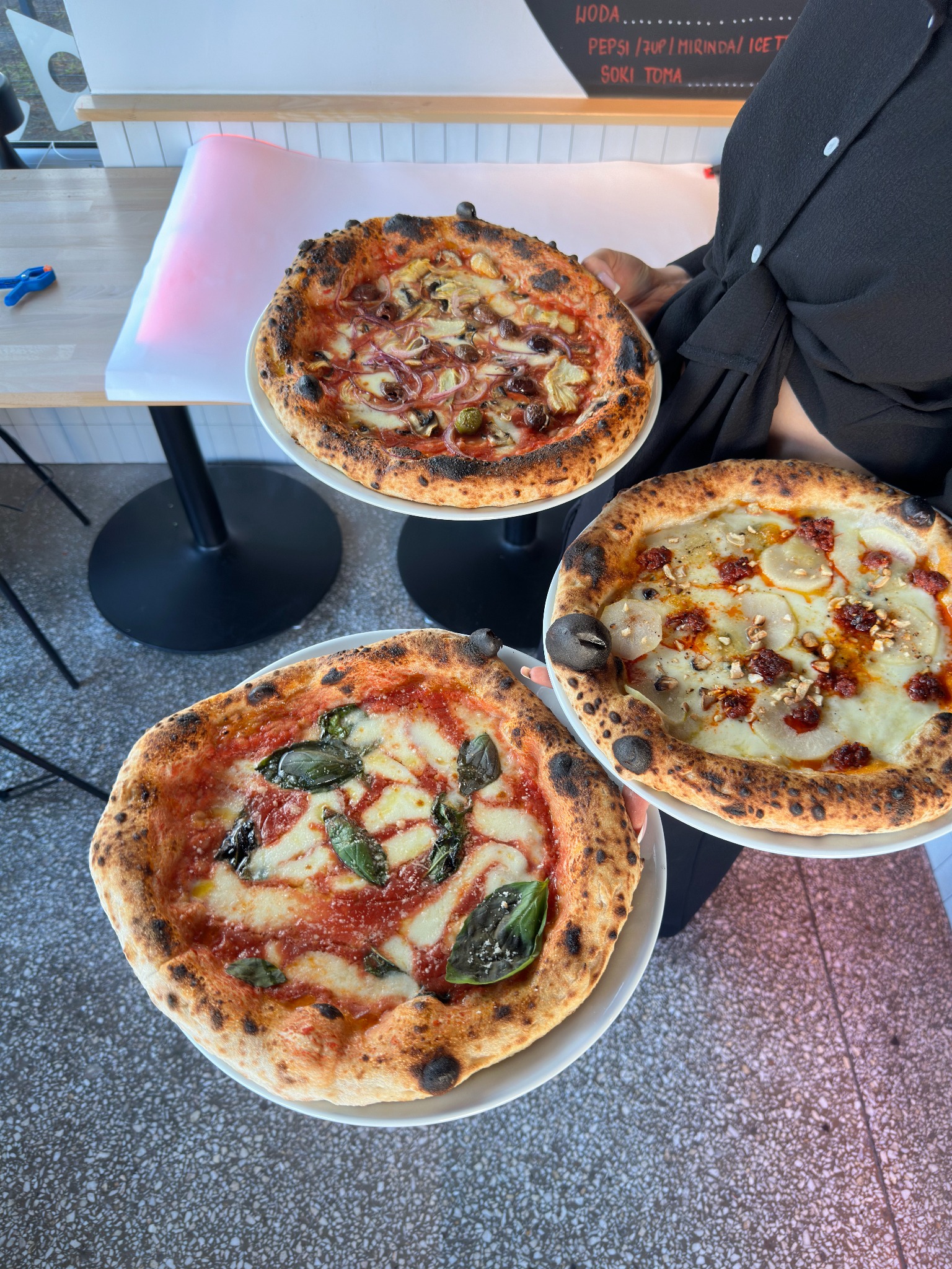 Trzy pizze na talerzach, każda z innym rodzajem dodatków, trzymane przez osobę w czarnej koszuli. Widoczne stoliki i menu na ścianie w tle. Jasne, naturalne oświetlenie.