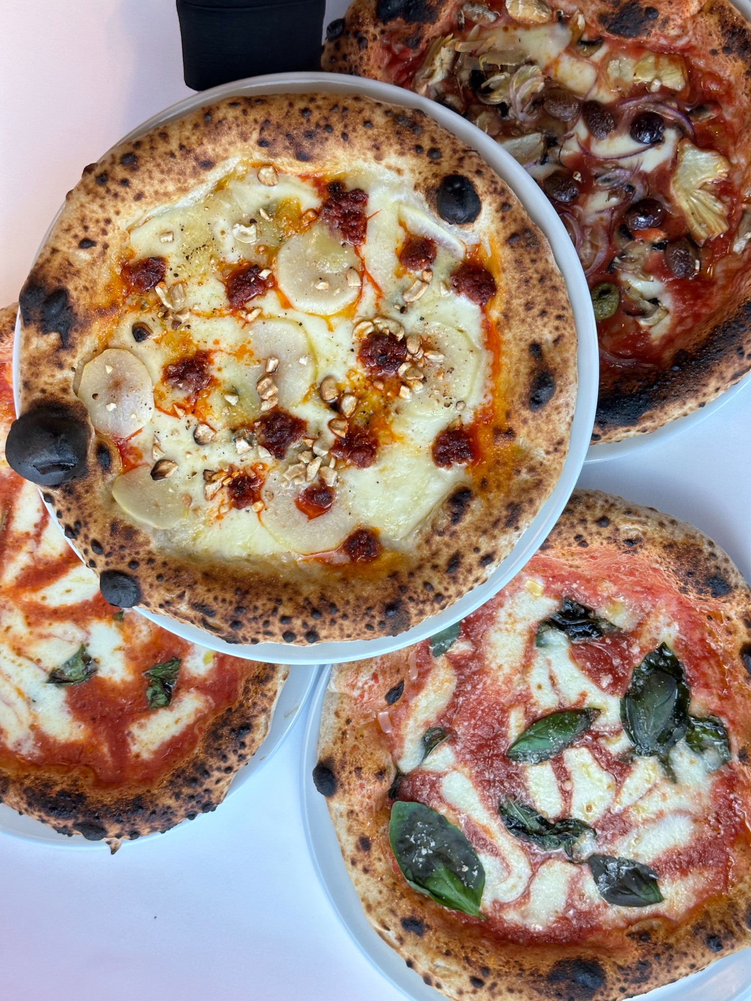 Cztery pizze na talerzach, każda z innym rodzajem dodatków, ułożone blisko siebie na białym tle. Widoczne przypalone brzegi ciasta i różnorodność składników.