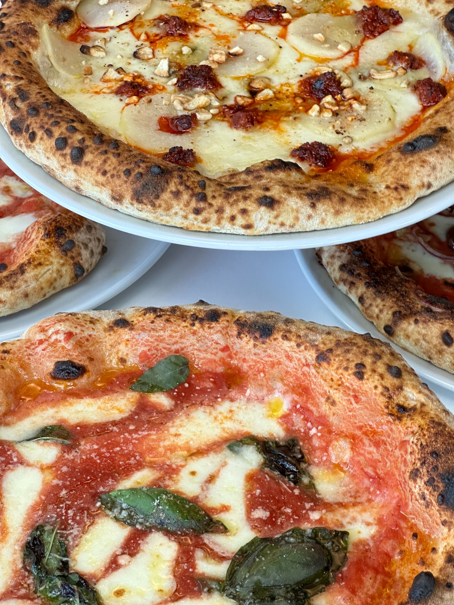 Dwie pizze na talerzach: jedna z sosem pomidorowym i bazylią, druga z serem, gruszką i suszonymi pomidorami. Widoczne przypalone brzegi ciasta.