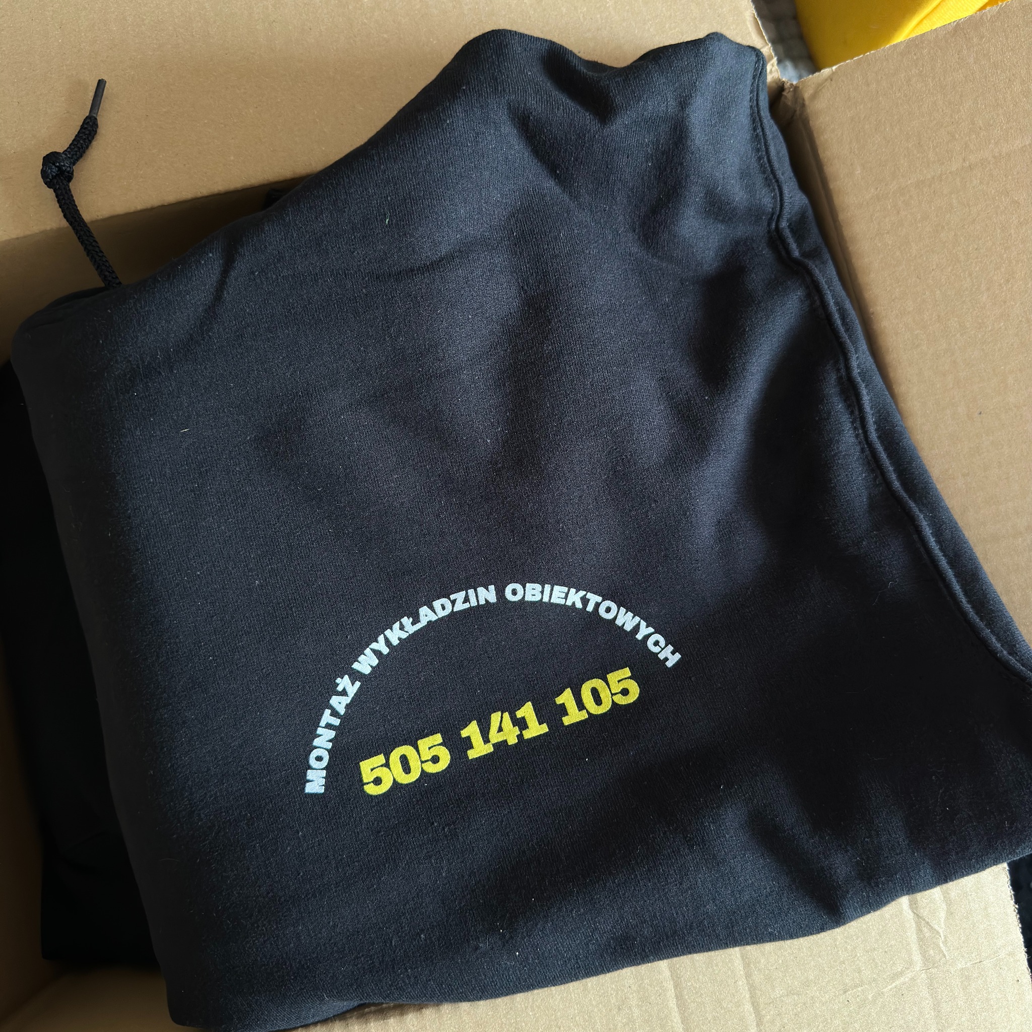 Czarna torba z nadrukiem 'Montaż wykładzin obiektowych' i numerem telefonu 505 141 105, leżąca w kartonowym pudełku. Widoczne sznurki ściągające torbę.