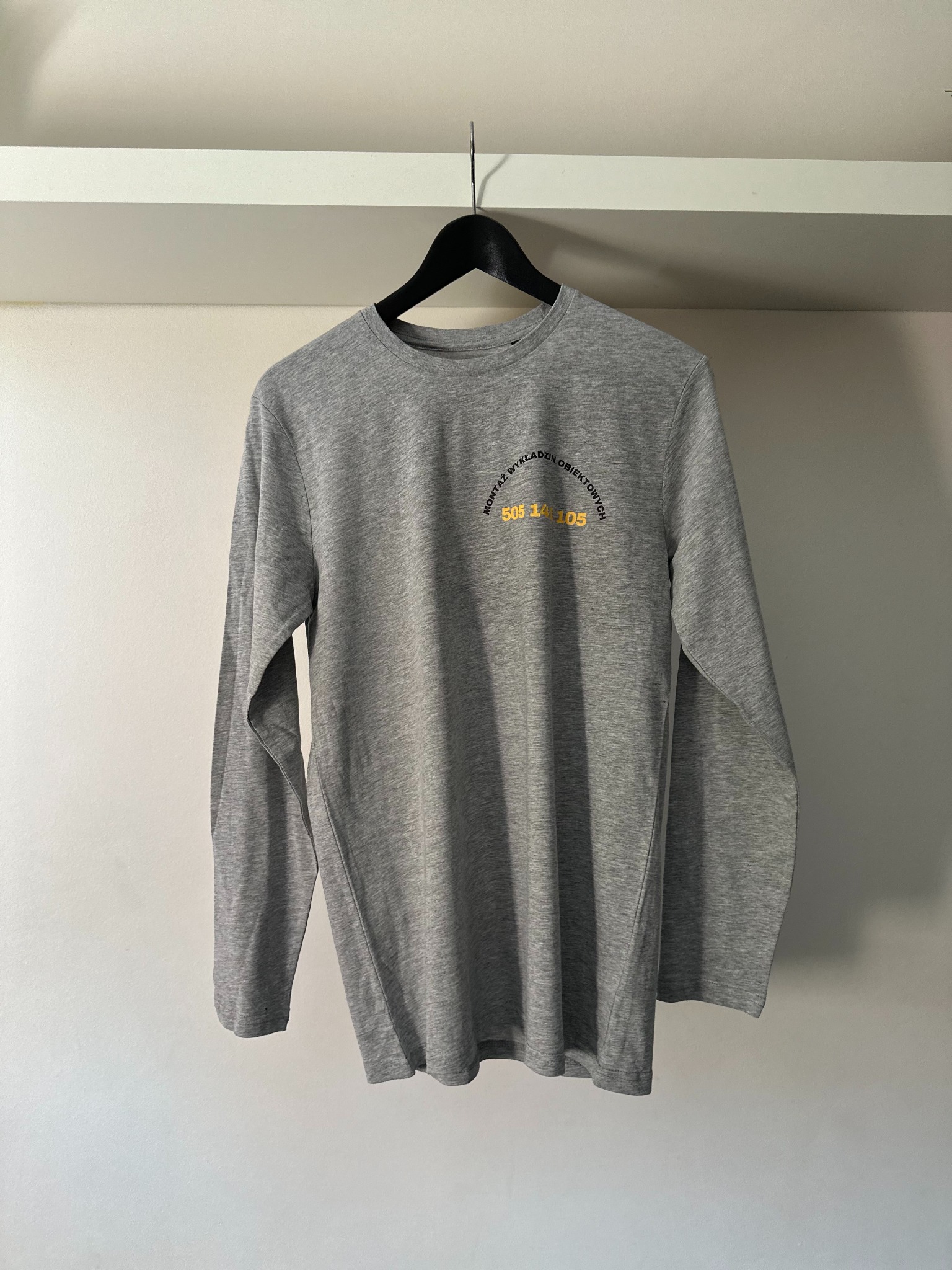 Szara koszulka z długim rękawem, wisząca na czarnym wieszaku. Na koszulce widnieje żółty nadruk z napisem 'Montaż wykładzin obiektowych' i numerem telefonu.