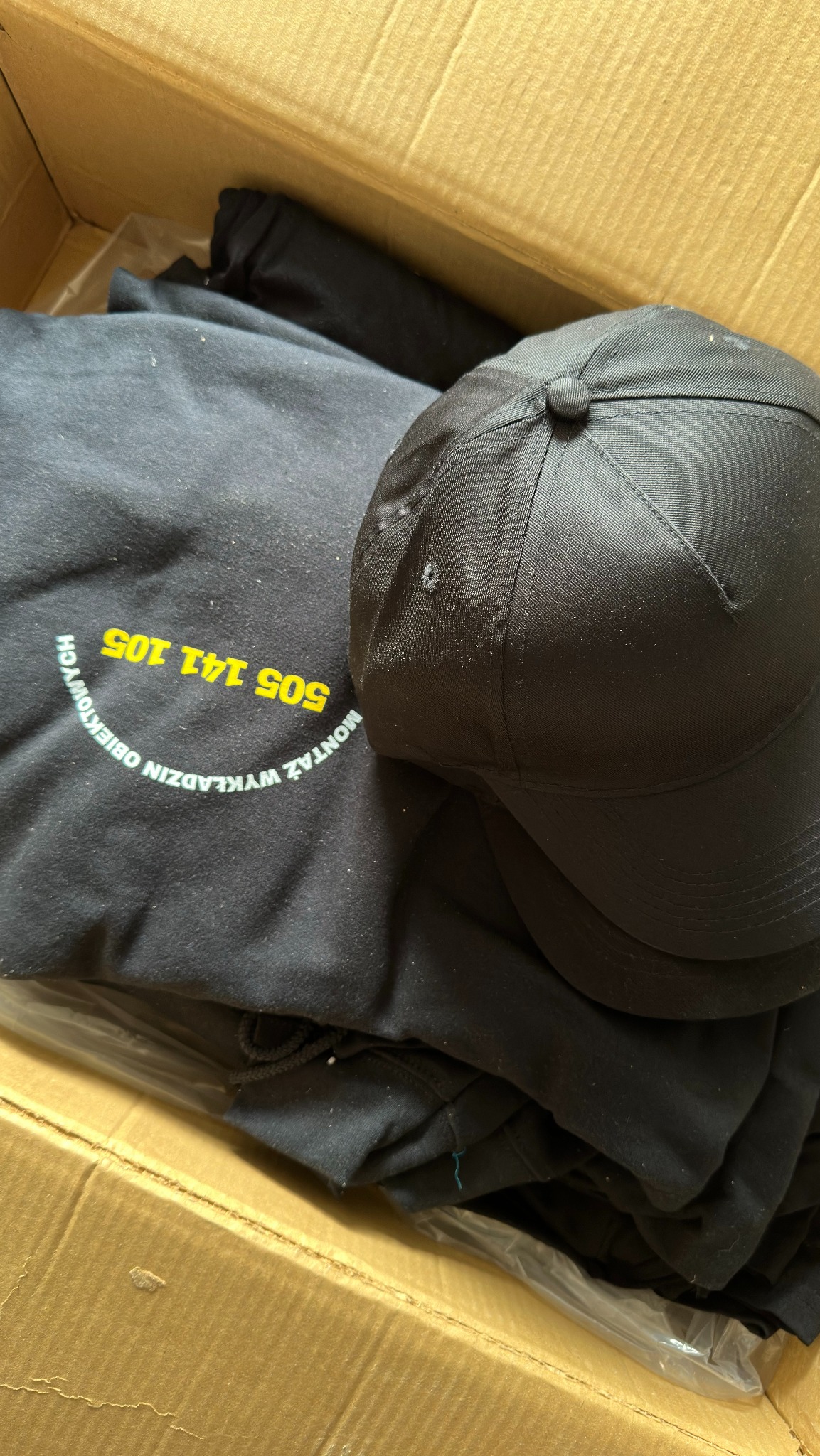 Czarna czapka i bluza z nadrukiem 'Montaż Wykładzin Obiektowych 505 141 105' w kartonowym pudełku. Widok z góry, oświetlenie naturalne.