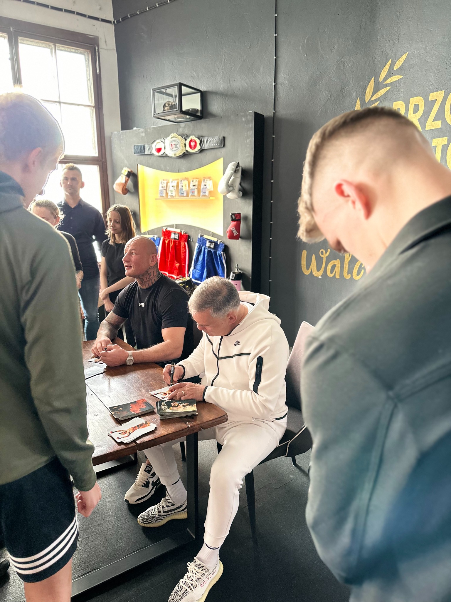 Dwóch mężczyzn podpisuje zdjęcia przy drewnianym stole w pomieszczeniu z bokserskimi akcentami i widzami. Jeden z nich ma tatuaże na szyi i rękach.