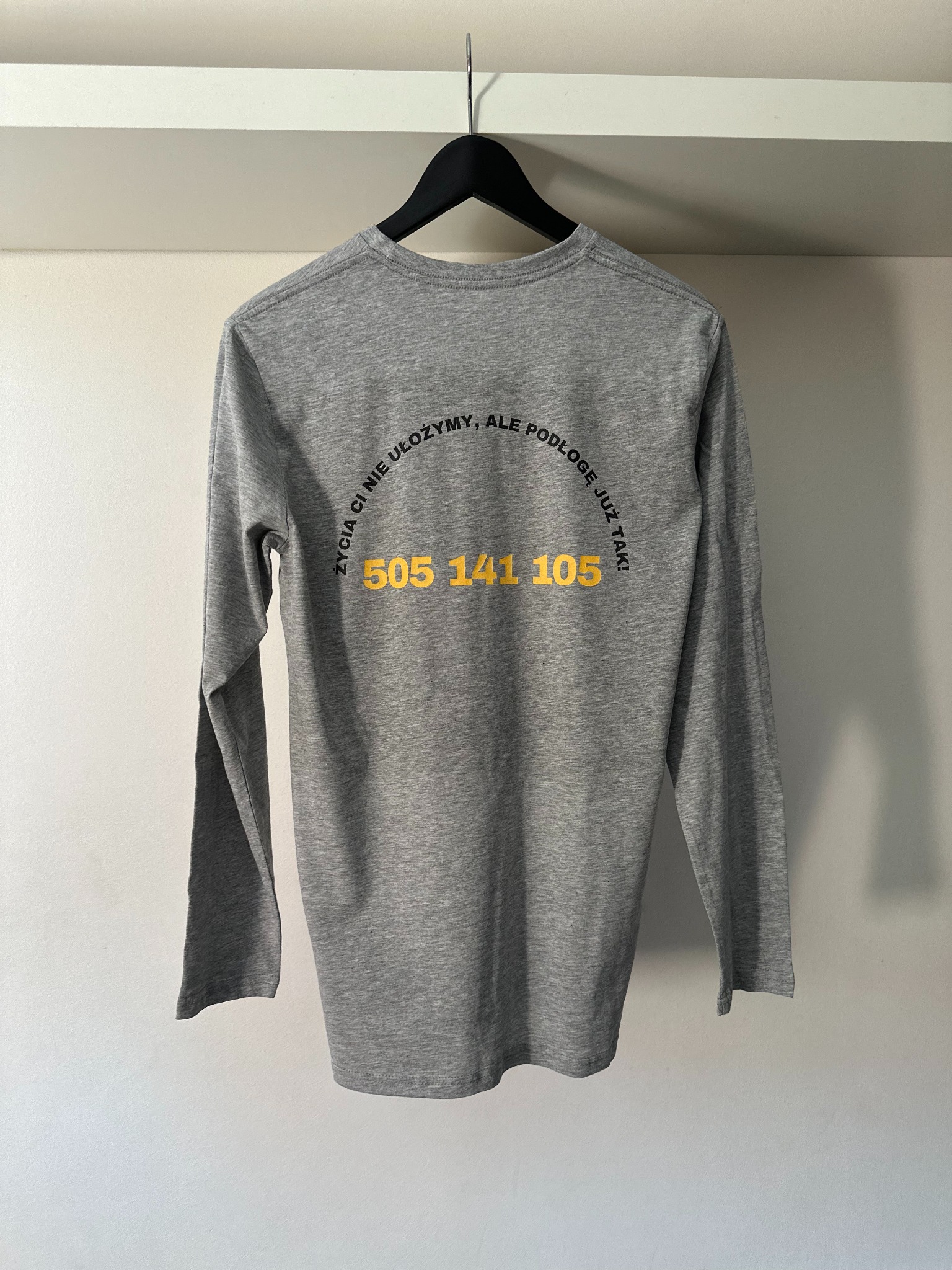 Szara koszulka z długim rękawem wisząca na wieszaku. Na koszulce nadruk z hasłem 'Życia Ci nie ułożymy, ale podłogę już tak!' i numerem telefonu w kolorze żółtym.