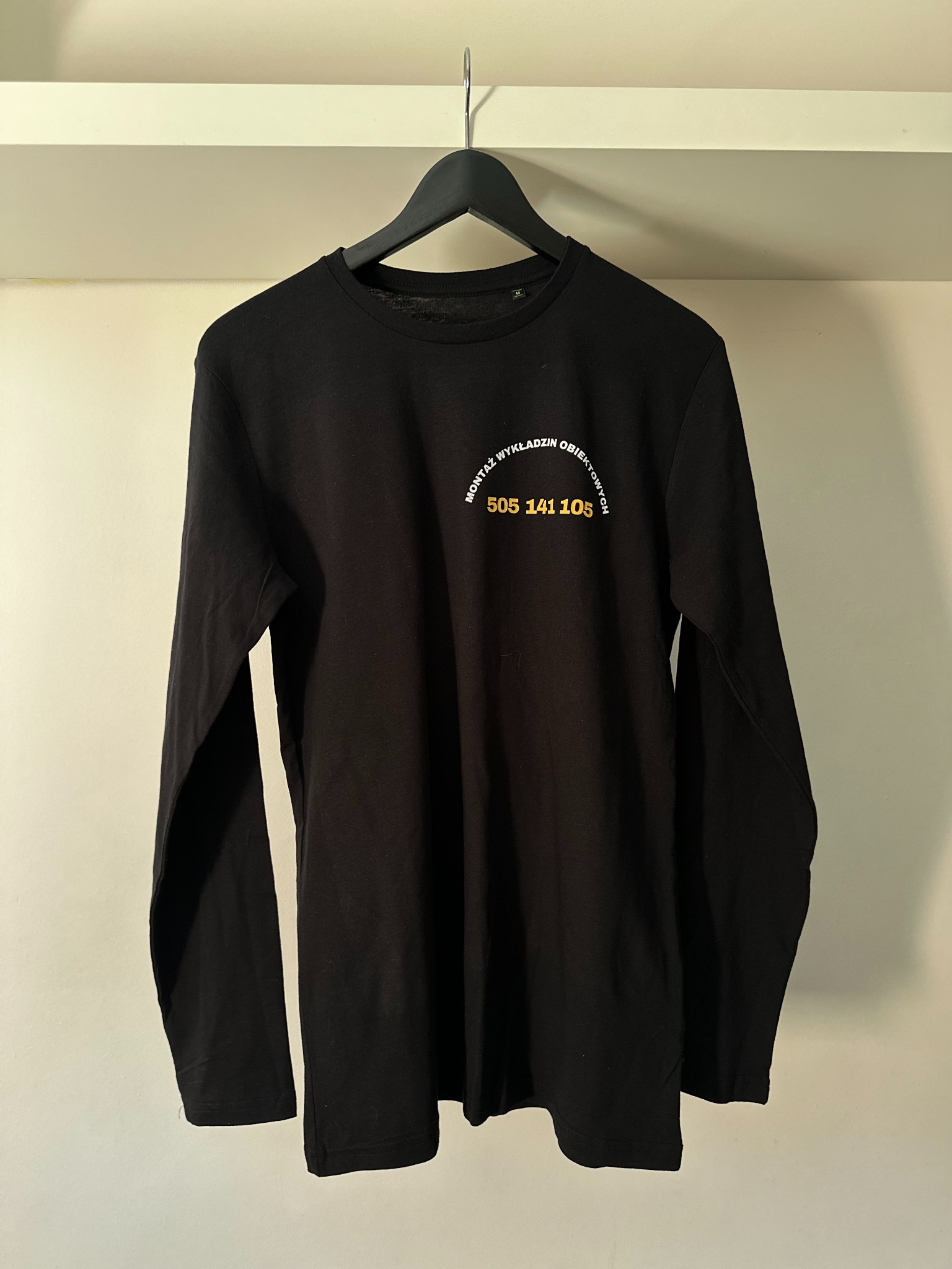 Czarna koszulka z długim rękawem na wieszaku, z nadrukiem 'Montaż wykładzin obiektowych' i numerem telefonu 505 141 105 w kolorze białym i żółtym.