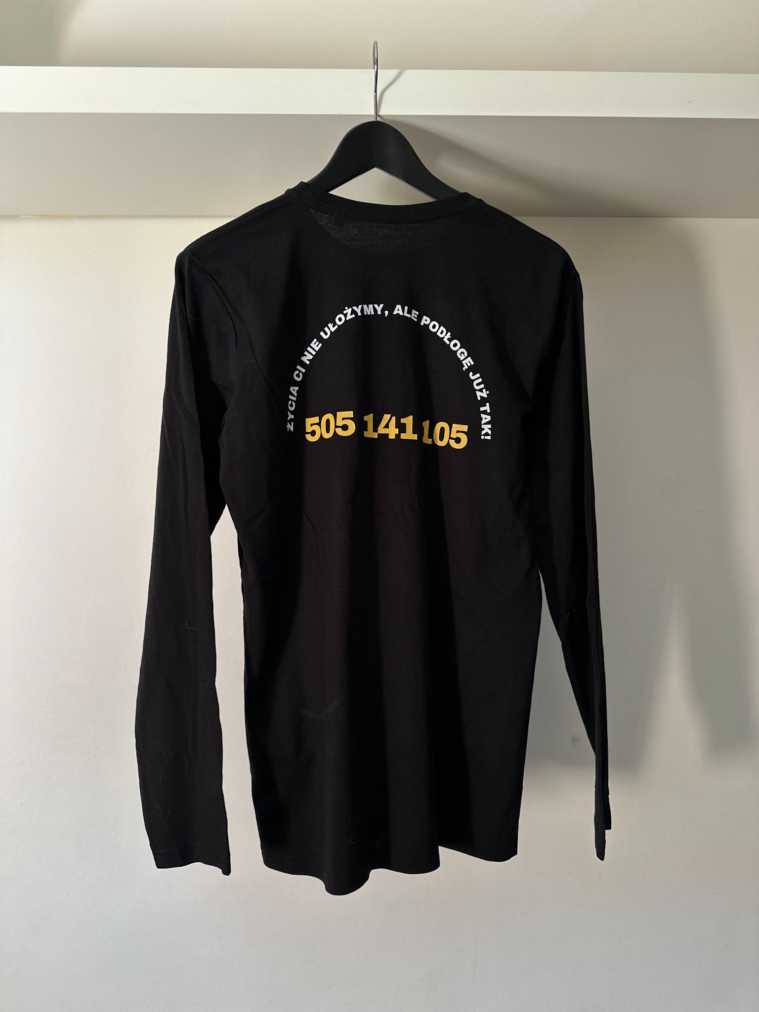 Czarna koszulka z długim rękawem, na wieszaku, z napisem 'Życia Ci nie ułożymy, ale podłogę już tak! 505 141 105' w kolorze białym i złotym.