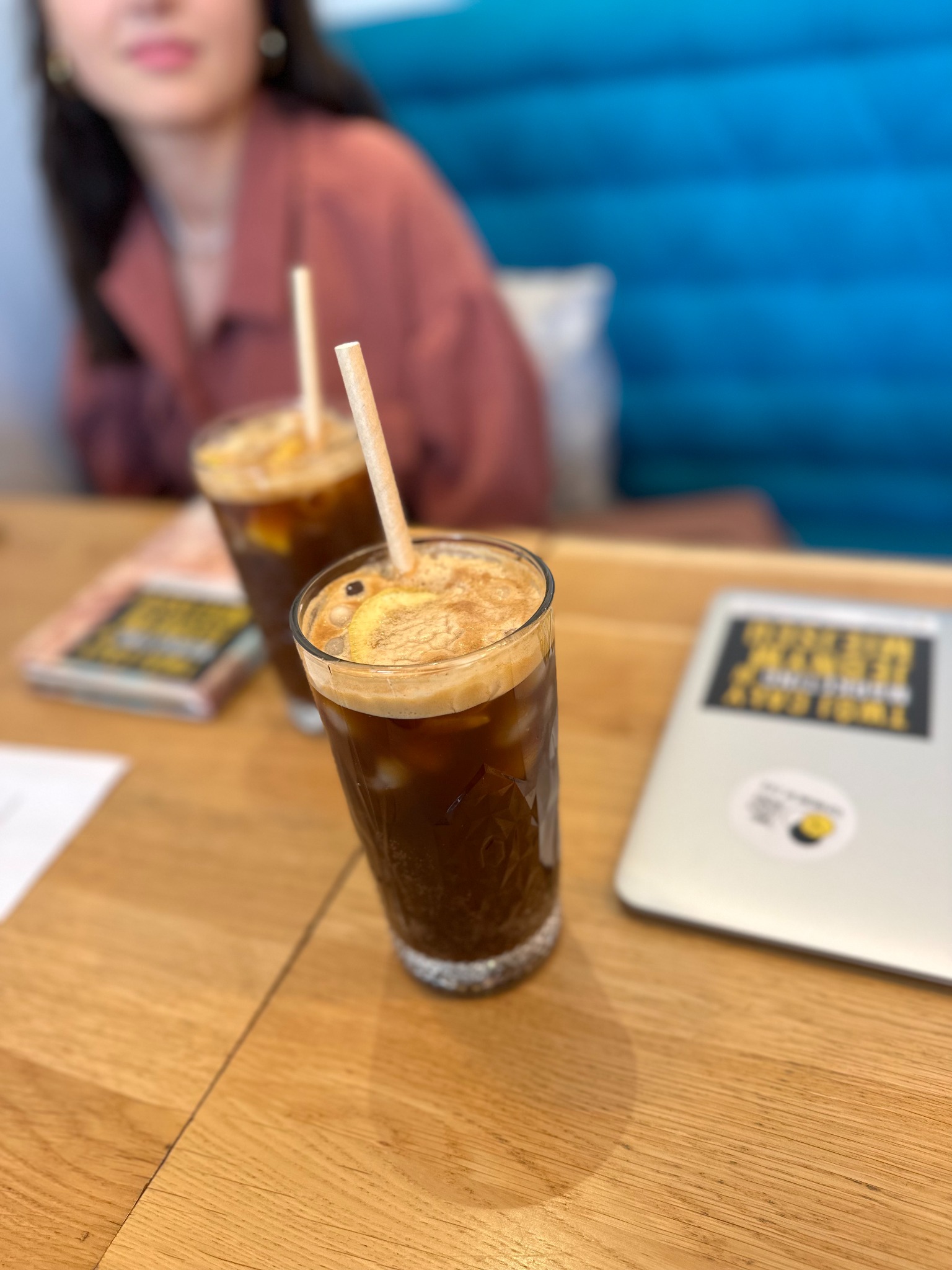 Dwa drinki z lodem i cytryną na drewnianym stole obok laptopa z naklejką. W tle rozmyta postać kobiety i niebieska kanapa. Ujęcie z bliska, naturalne światło.