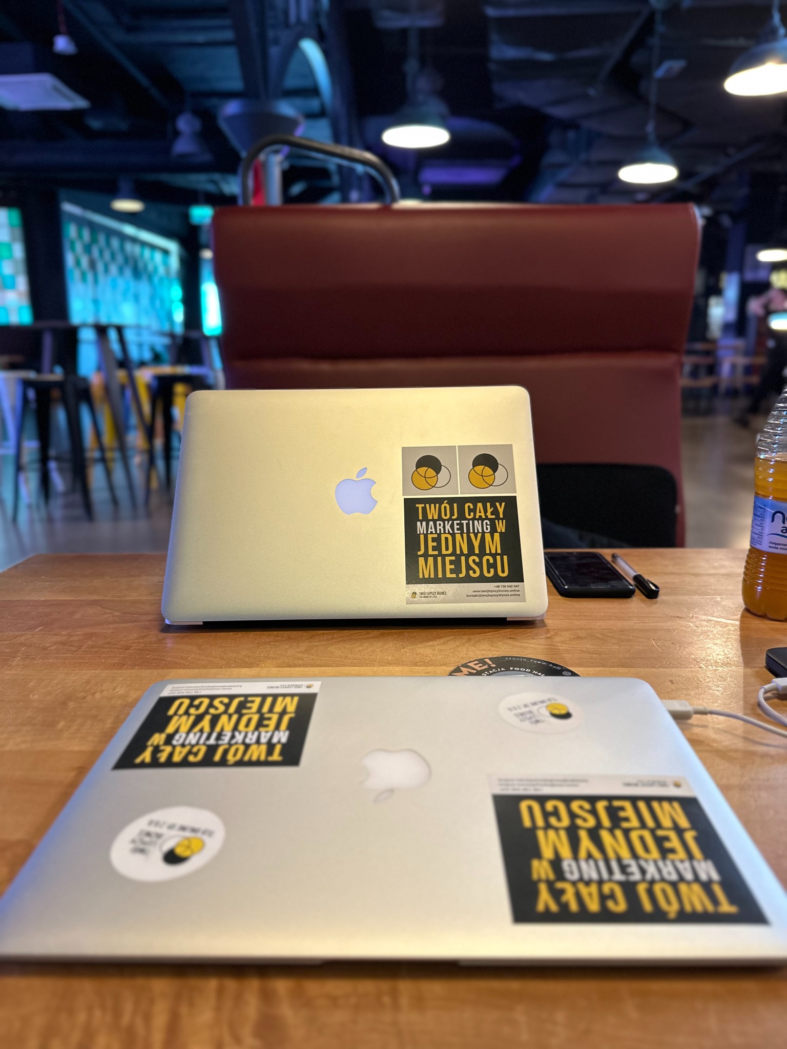 Dwa laptopy z naklejkami 'Twój cały marketing w jednym miejscu' na drewnianym stole w kawiarni. Widoczne logo Apple i butelka napoju. Ujęcie z bliska.