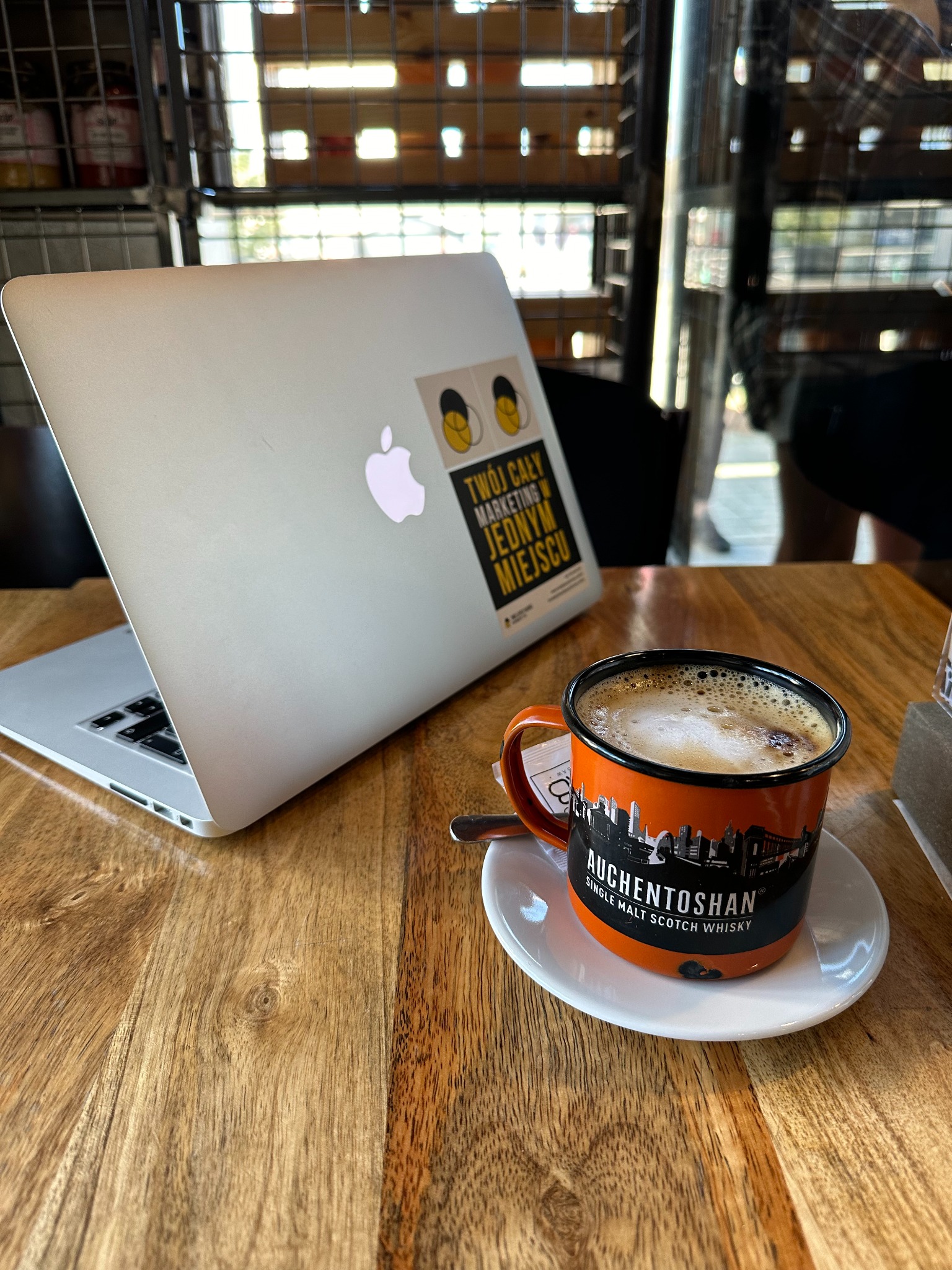 Laptop z naklejką marketingową i kubek kawy Auchentoshan na drewnianym stole, w tle metalowa konstrukcja. Praca zdalna w kawiarni.