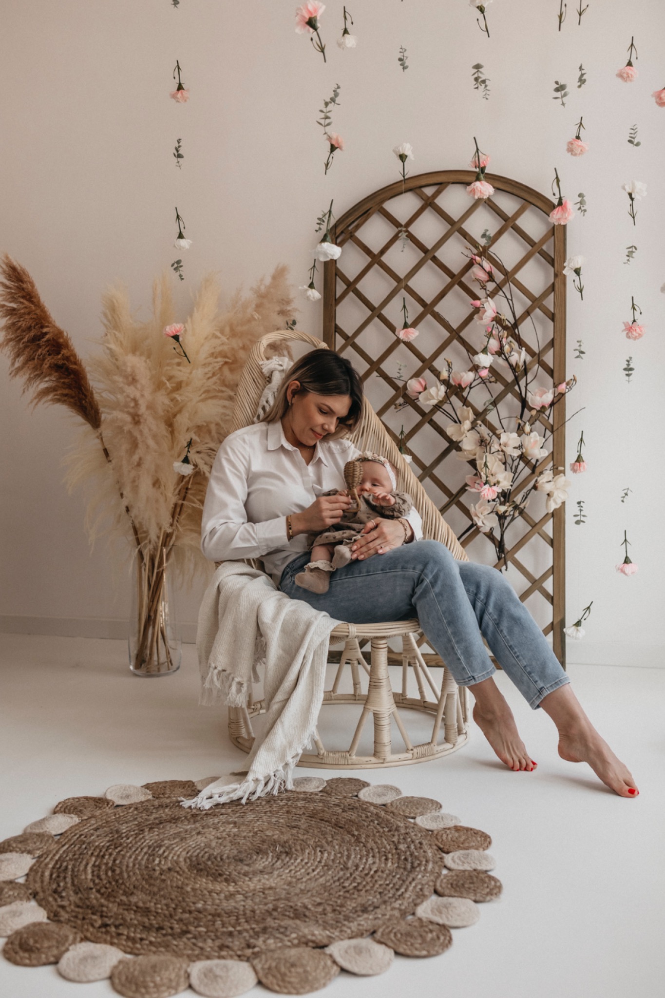 Kobieta w białej koszuli i jeansach siedzi w rattanowym fotelu, trzymając niemowlę w brązowej sukience i czapeczce, na tle ściany z kwiatowymi dekoracjami i pampasową trawą w wazonie.