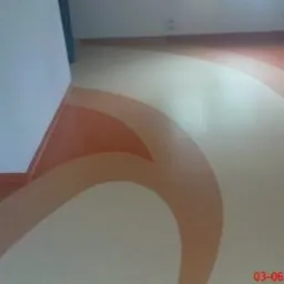 Wzorzysta podłoga z wykładziny PCV w odcieniach beżu i pomarańczy, z dekoracyjnymi łukami i pasami, wykończona przy ścianach listwami przypodłogowymi w kolorze podłogi.
