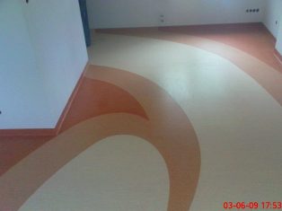 Wzorzysta podłoga z wykładziny PCV w odcieniach beżu i pomarańczy, z dekoracyjnymi łukami i pasami, wykończona przy ścianach listwami przypodłogowymi w kolorze podłogi.