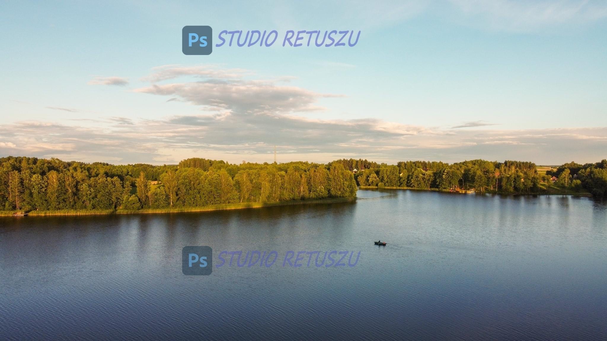 Panoramiczny widok z lotu ptaka na jezioro z łodzią, otoczone zielonym lasem w słoneczny dzień, z subtelnymi chmurami na niebie i logo studia retuszu nałożonym na obraz.