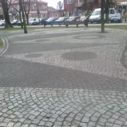 Plac miejski z geometrycznym wzorem ułożonym z szarej kostki brukowej, drzewa rosnące przez środek placu, w tle budynki i zaparkowane samochody.