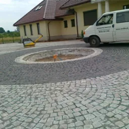 Układanie kostki granitowej w okrągły wz&oacute;r na podjeździe z centralnym klombem, widoczny samoch&oacute;d dostawczy firmy brukarskiej i ubijarka w tle.