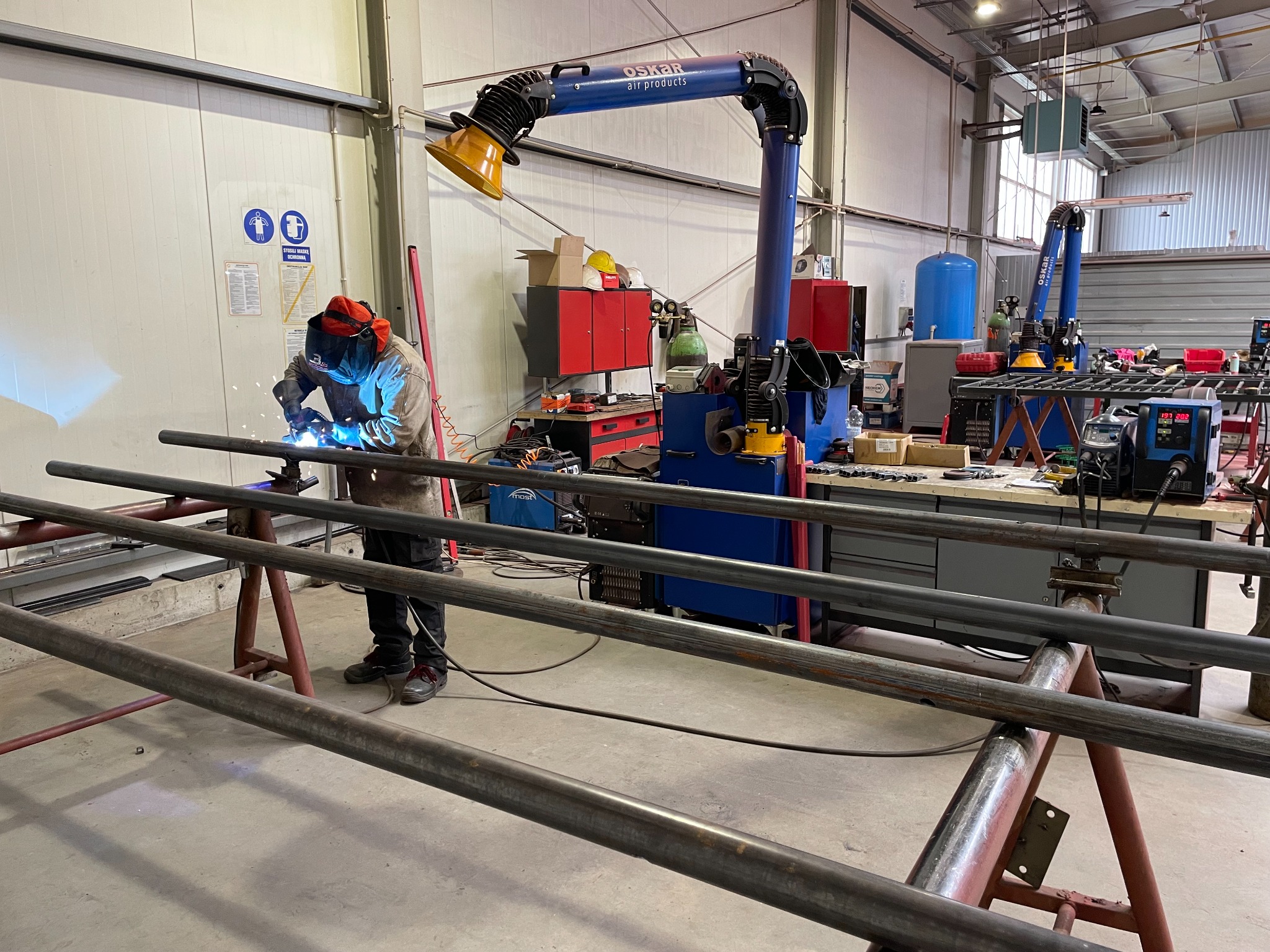 Spawanie metalowych rur w hali produkcyjnej z użyciem maski spawalniczej i odciągu dymu; widoczne iskry i sprzęt spawalniczy.